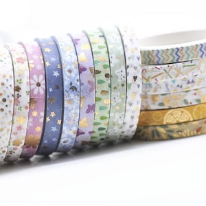 Op de afbeelding: Een verzameling decoratieve washi-tapes in verschillende kleuren en patronen. Ontwerpen omvatten bloemen, sterren, vlinders en geometrische motieven met goudfolie accenten. De tapes zijn gerangschikt in rollen en tonen een verscheidenheid aan stijlen voor knutselen en decoratie.