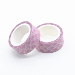 May include: Two rolls of decorative washi tape. The tape has a plaid pattern in shades of pink and white. The rolls are positioned on a white surface, with one slightly angled towards the viewer.
