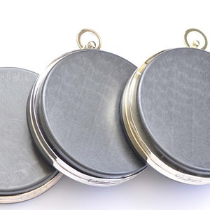 May include: Three round, silver and gold compacts with a dark gray, textured surface. Each compact has a small, round, gold loop on top.