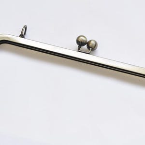 Könnte beinhalten: Ein rechteckiger Metall-Geldbörsenrahmen mit Vintage-Bronze-Finish. Der Rahmen hat einen Doppel-Kugelverschluss und kleine Schlaufen zum Befestigen an einer Tasche. Der Rahmen ist etwa 15 cm breit.