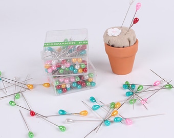 May include: A set of colorful glass-headed sewing pins in a clear plastic container. The pins are arranged in a variety of colors, including red, green, blue, and pink. The container is labeled "Dressmaker Pin".