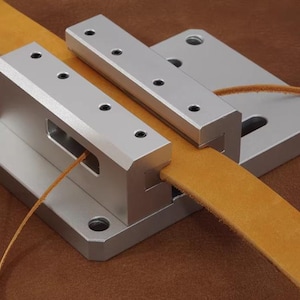 May include: A silver-colored leatherworking tool with a tan leather strap running through it. The tool has a rectangular base and a series of holes. The leather strap is a light brown color.
