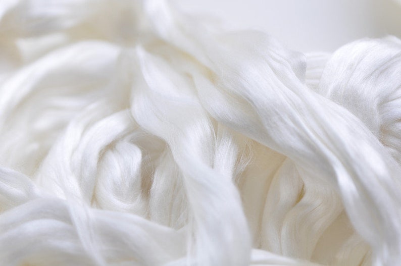 May include: Close-up of a pile of white silk fibers. The fibers are soft and flowing, with a subtle sheen. The image highlights the texture and delicate nature of the material, suggesting its use in textiles or crafting.