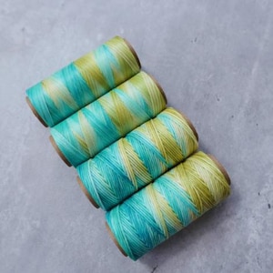 May include: Four spools of colorful thread. Each spool is wrapped with a gradient of teal and yellow thread. The spools are arranged diagonally on a gray surface. The thread appears to be for crafting or sewing.