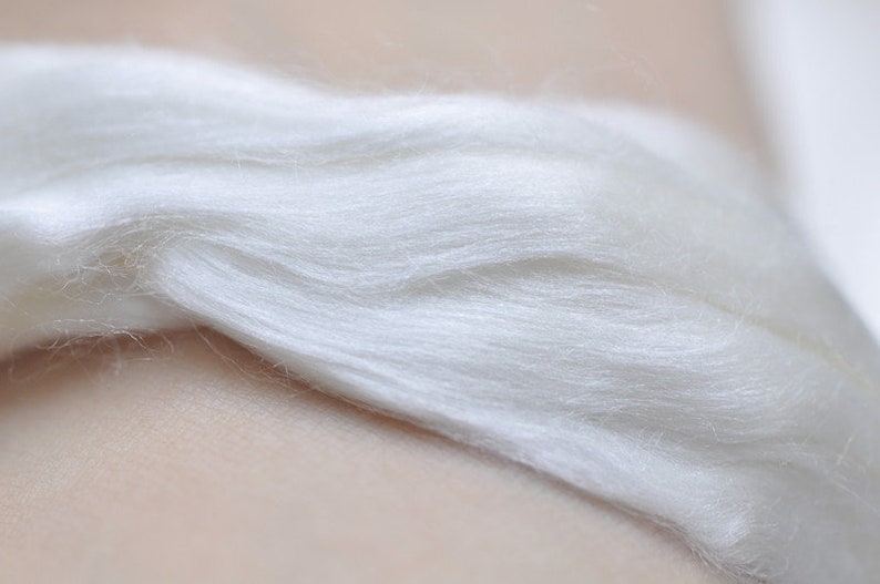 May include: Close-up of a bundle of soft, white fiber. The fibers are layered and appear to be smooth and delicate, with a slight sheen. The texture suggests a natural material, possibly silk or wool.