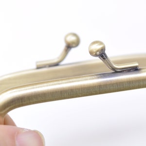 May include: Antique brass purse frame with two clasps. The frame is rectangular and has a smooth, brushed finish.