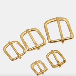 Puede incluir: Cinco hebillas de cinturón de metal dorado de diferentes tamaños. Las hebillas son todas rectangulares con una sola barra horizontal centrada.