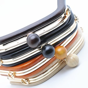 Puede incluir: Una pila de seis marcos de bolso de metal y madera de diferentes colores. Los marcos son todos del mismo tamaño y forma, pero tienen diferentes acabados. El marco superior es de madera marrón, el segundo es negro, el tercero es dorado, el cuarto es naranja, el quinto es beige y el sexto es de madera natural.