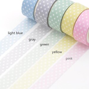 May include: A selection of washi tape rolls in light blue, grey, green, yellow, and pink. Each roll has a polka dot design. The tape is unrolled, showing the colours and patterns.