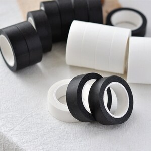 Puede incluir: Primer plano de rollos de cinta washi negra y blanca. Los rollos est&aacute;n apilados y dispuestos sobre una superficie blanca.