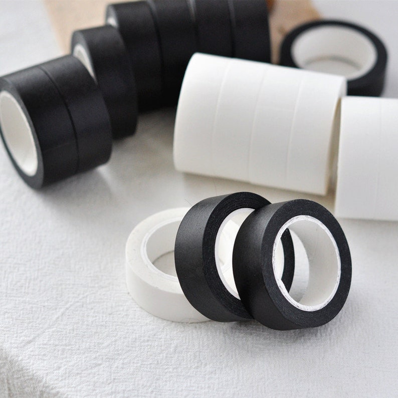 Puede incluir: Primer plano de rollos de cinta washi negra y blanca. Los rollos est&aacute;n apilados y dispuestos sobre una superficie blanca.