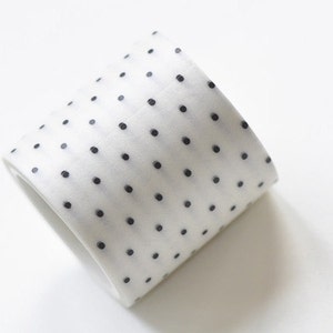 May include: A roll of white washi tape featuring a pattern of small, black polka dots. The tape is angled, showing its rolled shape. The background is plain white.