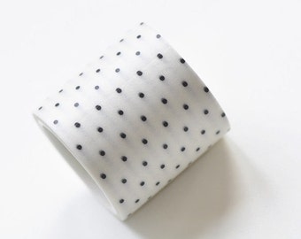May include: A roll of white washi tape with a repeating pattern of small, evenly spaced black dots. The tape is slightly angled, revealing its rolled form. The background is plain white.