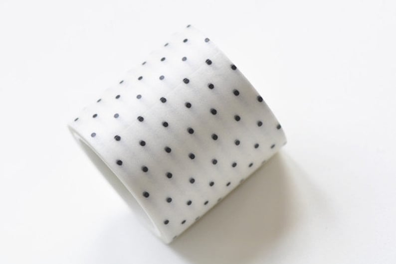 May include: A roll of white washi tape with a repeating pattern of small, evenly spaced black dots. The tape is slightly angled, revealing its rolled form. The background is plain white.