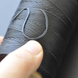 Può includere: Primo piano di una bobina di filo nero. Il filo è avvolto strettamente, con un anello estratto. Il filo sembra resistente, forse cerato, e adatto per lavori di artigianato o gioielleria.