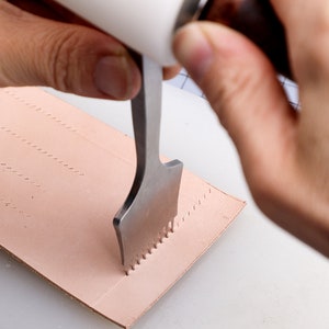 May include: A hand uses a leather stitching tool to punch holes in a piece of light brown leather. The tool has a white handle and a metal head with multiple needles.