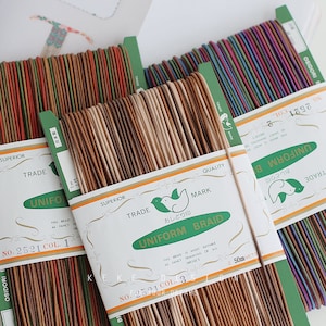 May include: Spools of colorful uniform braid in various shades of brown, green, red, and purple. Each spool is labeled with text and a bird logo. The braid is suitable for fancy trimming of dresses.