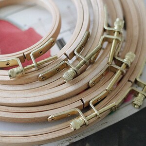 Puede incluir: Un conjunto de aros de bordado de madera de varios tamaños, apilados uno encima del otro. Los aros son de madera natural y tienen mecanismos de apriete de tornillo de latón.