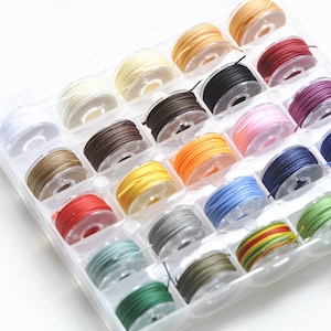 Puede incluir: Un organizador de plástico transparente lleno de carretes de hilo de colores. Los colores del hilo incluyen blanco, crema, marrón, negro, rosa, rojo, naranja, amarillo, azul y verde. Los carretes están ordenados en compartimentos.