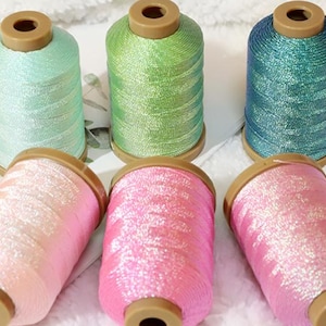 May include: Six spools of colorful thread in shades of mint green, light green, and teal, along with pink and rose. Each spool has a brown top and base. The threads appear to have a slight shimmer.