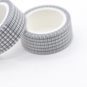 May include: Two rolls of decorative washi tape. The tape is grey with a grid pattern and a white interior. The rolls are positioned on a white background. This tape is ideal for scrapbooking, journaling, and crafting projects.