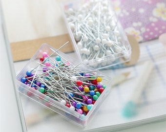 May include: Two clear plastic containers filled with sewing pins. One container has white pearl-headed pins, the other has multi-colored plastic-headed pins.