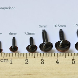 Puede incluir: Comparaci&oacute;n de tama&ntilde;o de seis ojos de seguridad de pl&aacute;stico negro con iris marrones, que van desde 4,5 mm hasta 12 mm. Los ojos est&aacute;n alineados en una regla de madera con marcas de cent&iacute;metros.