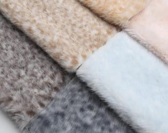 May include: Close-up of various faux fur fabric swatches in shades of beige, gray, and blue. The fabrics have a soft, textured appearance, with visible fibers and a plush feel. The arrangement showcases the different colors and textures.