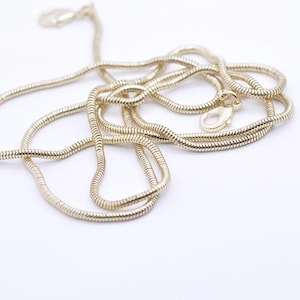 May include: A gold-coloured snake chain necklace with a lobster clasp closure. The chain is coiled and lies on a white surface. The necklace is a simple, elegant piece of jewellery.