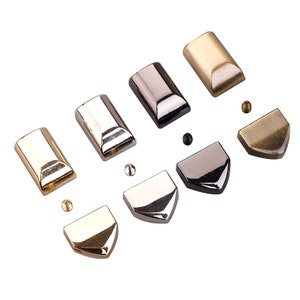 Könnte beinhalten: Satz mit sechs Metallteilen für Taschenzubehör in Gold, Silber und Antikmessing. Die Teile umfassen vier rechteckige Kappen und zwei spitze Kappen.
