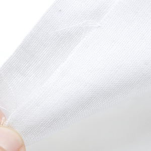 Può includere: Primo piano di un tessuto bianco con una trama tessuta. Il tessuto è tenuto da una mano e i bordi sono sfilacciati.