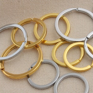 May include: A group of gold and silver key rings. The key rings are made of metal and have a smooth finish. They are arranged in a pile on a beige fabric background.