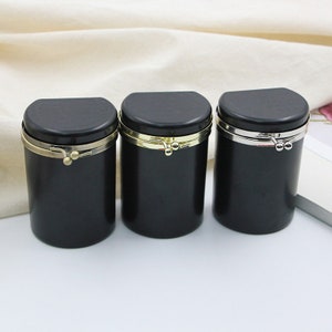 May include: Three black cylindrical containers with a hinged lid and a metal clasp. The clasps are gold, silver, and gold.