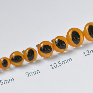 May include: A set of six orange plastic safety eyes with black pupils, ranging in size from 7.5 mm to 12 mm. The eyes are arranged in order of size, with the smallest on the left and the largest on the right.