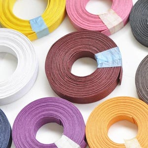 Può includere: Una collezione di strisce di carta colorate in varie tonalità, tra cui giallo, bianco, rosa, grigio, bordeaux, marrone, viola, arancione e blu navy. Ogni rotolo è fissato con un nastro adesivo, mostrando una varietà di forniture artigianali.