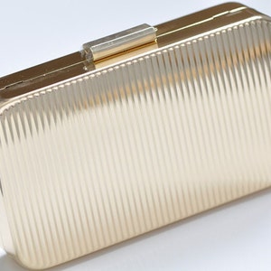 May include: A rectangular gold clutch with a ridged surface. The clutch has a gold clasp and a rectangular shape. The ridged texture creates a play of light and shadow on the surface. The overall design is sleek and elegant.