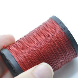 May include: A spool of red thread, held in a hand, against a white background. The thread is tightly wound on a black plastic spool. The spool has text printed on the side.
