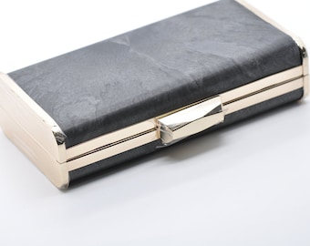 Può includere: Una pochette rettangolare con esterno grigio scuro strutturato e finiture in metallo color oro. La pochette ha una chiusura a scatto e un design elegante, adatta per eventi formali o per l'uso quotidiano.