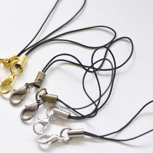 May include: Three black phone straps with different coloured metal clasps and end caps. The clasps are gold, silver, and bronze.
