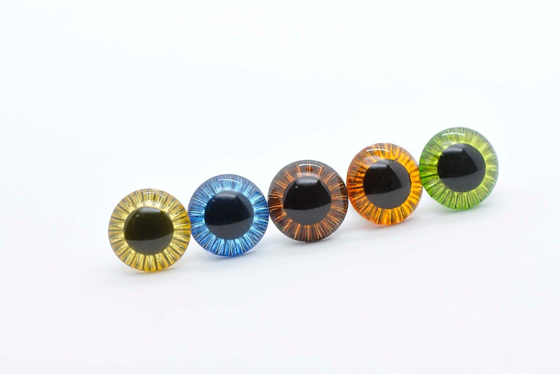 May include: Five different coloured plastic safety eyes with black pupils. The eyes are arranged in a row from left to right: yellow, blue, orange, amber, and green.