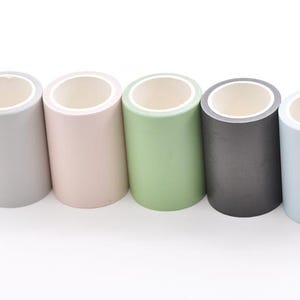 Könnte beinhalten: Fünf Rollen Washi-Tape in einer Reihe, jede in einer anderen Pastellfarbe. Die Farben sind Grau, Rosa, Grün, Dunkelgrau und Hellblau. Jede Rolle hat eine weiße Innenseite und steht aufrecht.
