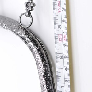 May include: A silver-toned metal purse frame with an ornate design and a flower-shaped clasp. The frame is 6 inches long and is measured against a ruler.