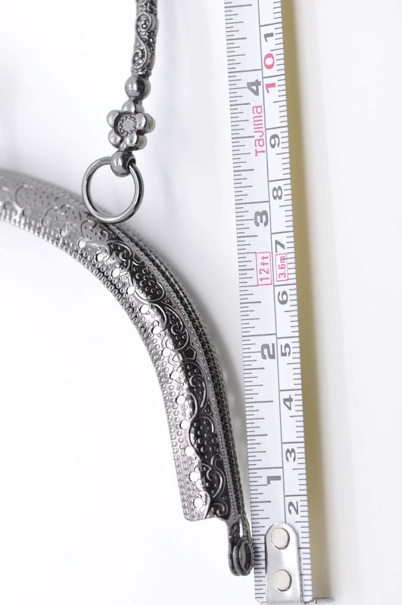 May include: A silver-toned metal purse frame with an ornate design and a flower-shaped clasp. The frame is 6 inches long and is measured against a ruler.