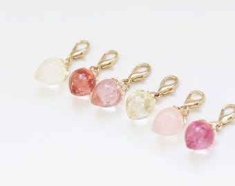 May include: A collection of six strawberry-shaped charms in varying shades of pink, red, and yellow. Each charm has a gold-colored clasp. The charms are arranged on a white surface.