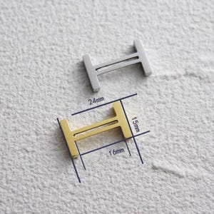 May include: Two metal H-shaped charms, one silver and one gold. The gold charm is 1.6cm wide and 1.5cm high, with a total length of 2.4cm. The charms are displayed on a textured, light-coloured surface.