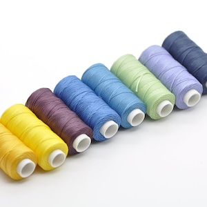 Peut inclure: Huit bobines de fil de différentes couleurs, dont le jaune, le violet, le bleu, le vert et le bleu clair. Les bobines sont disposées en rangée du plus foncé au plus clair.