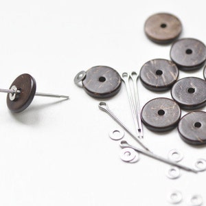 May include: A selection of dark brown, circular wooden beads with a central hole, accompanied by silver-coloured metal pins, washers, and fasteners. The beads display a natural wood grain. Suitable for jewellery making or craft projects.