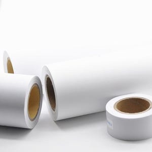 Puede incluir: Varios rollos de papel blanco se muestran sobre un fondo blanco. Los rollos varían en tamaño, con el más grande en el fondo y uno más pequeño en primer plano. El papel tiene un acabado liso y mate, y los núcleos son de color marrón claro.