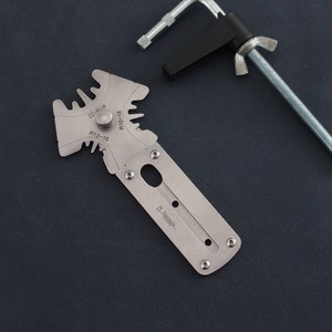 May include: A silver metal tool with a ruler and a series of different sized circles marked with measurements in inches. The tool is labeled "R12-16", "R16-18", and "R22-23". The tool is on a dark gray surface.