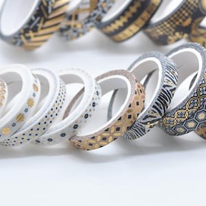 Puede incluir: Una colección de rollos de cinta washi decorativa con varios diseños. Los diseños incluyen flores doradas, lunares, formas geométricas y rayas de cebra. La cinta es blanca con detalles dorados, negros y plateados. Ideal para manualidades y scrapbooking.